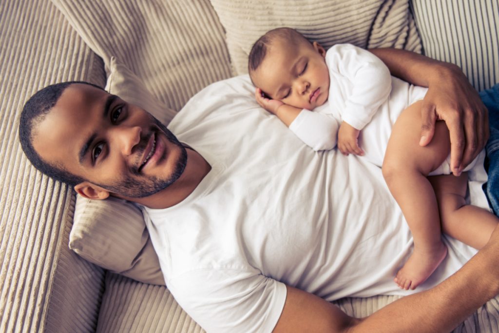 smiling man laying on couch holding sleeping baby.