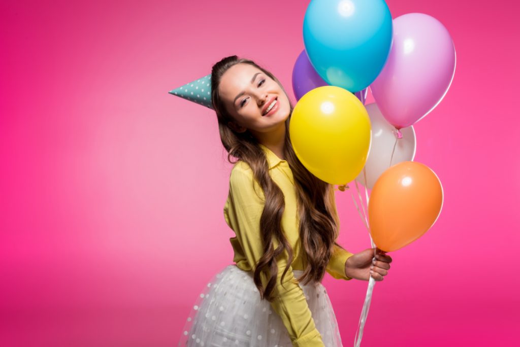 smiling young woman with blue birthday hat and holding colorful balloons.