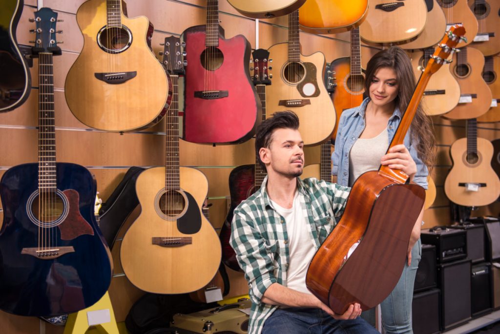 man holding wood guitar with woman and wall of guitars behind.