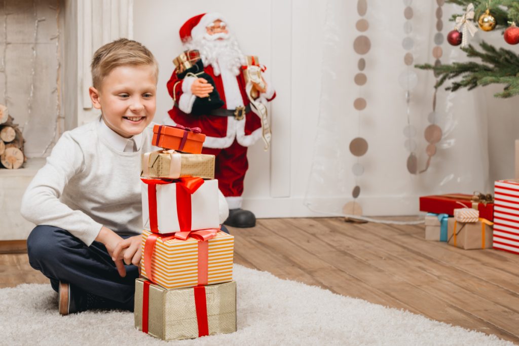 smiling child with stack of 5 wrapped gifts in front of Santa figure.
